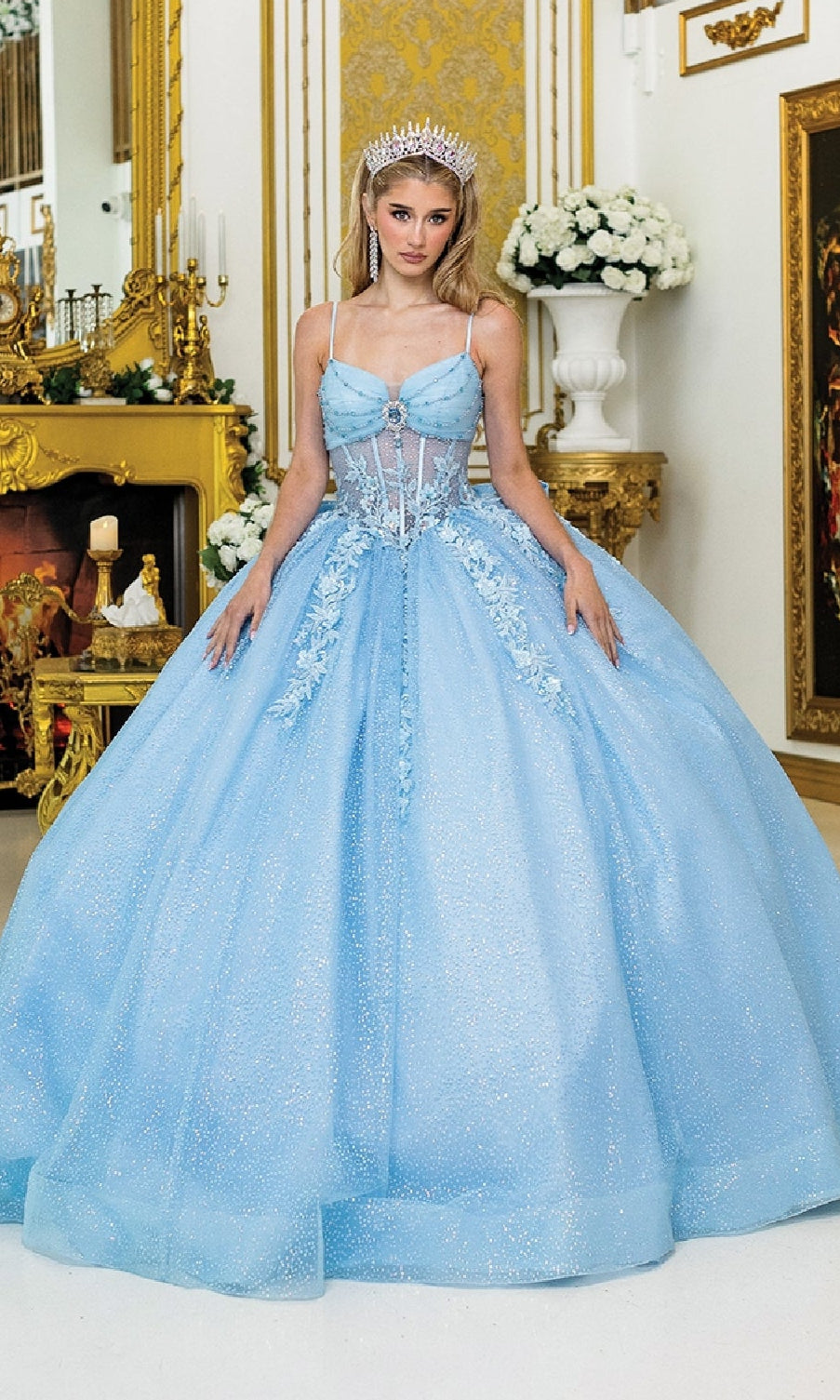 A woman in the Dancing Queen 1958 Long Quinceañera Dress with Bow stands in an ornate, gold-decorated room adorned with elegant floral arrangements, wearing a sparkling tiara.
